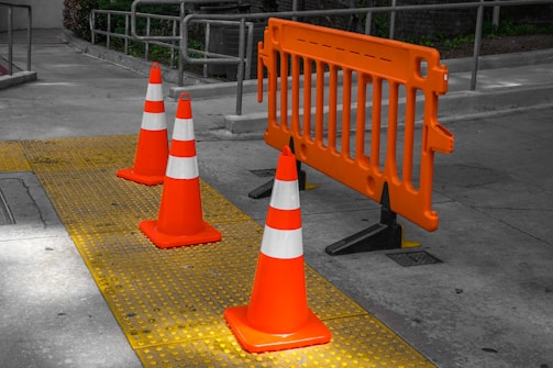 Orange traffic cones are lined up along a yellow tactile paving, accompanied by an orange safety barrier on a gray pavement. The scene suggests an area under construction or repair, cordoned off for safety.