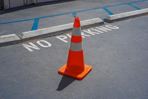 A bright orange traffic cone is placed on an asphalt surface with the words 'NO PARKING' painted in white. Blue lines and concrete barriers can be seen in the background, indicating a parking area.