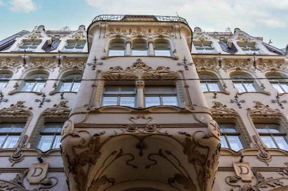 An ornate building facade featuring intricate, detailed carvings and embellishments around the windows and along the surface. The architecture displays an Art Nouveau style with floral and organic motifs. The upper levels include curved balconies and decorative elements.