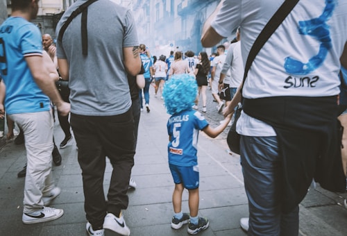 A group of people walking down a street, many wearing matching sports shirts. A young child adorned in a blue sports outfit with a blue wig stands out amongst the crowd. The atmosphere appears lively, possibly part of a sports event or parade.