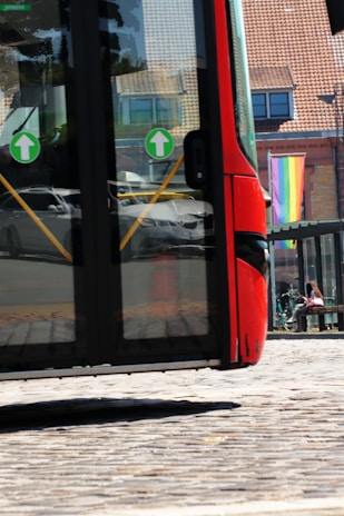 A partially visible red bus, with large glass windows showing reflections of cars and surroundings. Two green directional arrow stickers are on the windows. In the background, a multi-colored rainbow flag is hanging outside a brick building with multiple windows. A person sits on a bench near bicycles, suggesting an urban setting.