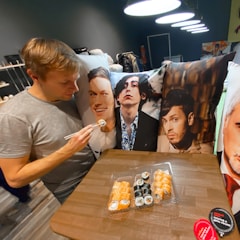 A person is using chopsticks to eat sushi in front of a table with several pillows featuring printed photographs of various individuals. The setting is indoors with modern lighting and a dark background.