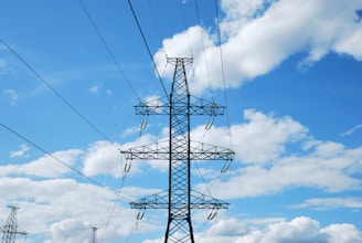 A metal electricity pylon stands tall against a bright blue sky scattered with white clouds. The pylon carries multiple high-voltage power lines that stretch across the image.