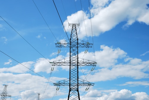A metal electricity pylon stands tall against a bright blue sky scattered with white clouds. The pylon carries multiple high-voltage power lines that stretch across the image.