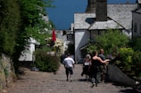 A quaint cobblestone street lined with lush greenery and flowers leads down to a seaside view. People are casually walking towards a traditional building with a sign that reads 'New Inn Hotel'. The scene is full of vibrant vegetation, with red and white flowers contrasting against the natural stone and timber buildings.