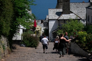 A quaint cobblestone street lined with lush greenery and flowers leads down to a seaside view. People are casually walking towards a traditional building with a sign that reads 'New Inn Hotel'. The scene is full of vibrant vegetation, with red and white flowers contrasting against the natural stone and timber buildings.