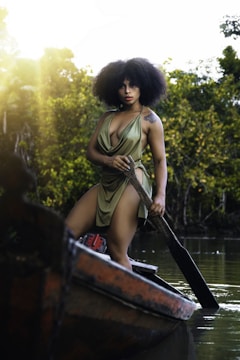 A person with voluminous curly hair is standing on a small boat in a lush, green waterway, holding a paddle. The person is wearing a flowing, green dress with a deep neckline. The background features dense trees and foliage, with sunlight filtering through the leaves.