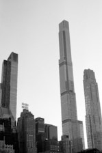 A monochromatic photograph featuring a skyline of modern skyscrapers with geometric designs. The tallest building rises prominently at the center, surrounded by a mix of other high-rise buildings. The iconic Essex House sign is visible on one of the buildings.