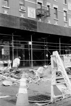 Two construction workers are engaged in roadwork, surrounded by rubble and debris in a city environment. Scaffolding lines the building behind them, with signs indicating ongoing repairs. Traffic cones and barriers are positioned around the work area.