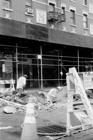 Two construction workers are engaged in roadwork, surrounded by rubble and debris in a city environment. Scaffolding lines the building behind them, with signs indicating ongoing repairs. Traffic cones and barriers are positioned around the work area.