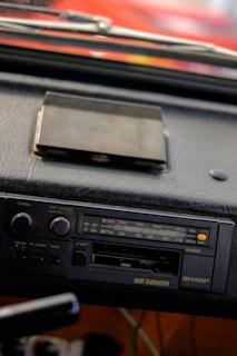 A close-up view of a vintage car stereo system mounted on a dashboard. The stereo features an AM/FM radio tuner, volume and tuning knobs, and a cassette tape slot. There's an old cassette tape case placed on the dashboard above the stereo. The dashboard appears to be made of black leather material.