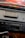 A close-up view of a vintage car stereo system mounted on a dashboard. The stereo features an AM/FM radio tuner, volume and tuning knobs, and a cassette tape slot. There's an old cassette tape case placed on the dashboard above the stereo. The dashboard appears to be made of black leather material.