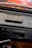 A close-up view of a vintage car stereo system mounted on a dashboard. The stereo features an AM/FM radio tuner, volume and tuning knobs, and a cassette tape slot. There's an old cassette tape case placed on the dashboard above the stereo. The dashboard appears to be made of black leather material.