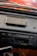 A close-up view of a vintage car stereo system mounted on a dashboard. The stereo features an AM/FM radio tuner, volume and tuning knobs, and a cassette tape slot. There's an old cassette tape case placed on the dashboard above the stereo. The dashboard appears to be made of black leather material.
