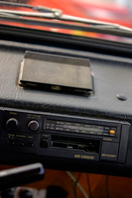 A close-up view of a vintage car stereo system mounted on a dashboard. The stereo features an AM/FM radio tuner, volume and tuning knobs, and a cassette tape slot. There's an old cassette tape case placed on the dashboard above the stereo. The dashboard appears to be made of black leather material.