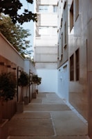 A narrow courtyard lined with small trees in rectangular planters is bordered by tall buildings. The space features a modern, minimalist design with large stone tiles on the ground and minimal decoration. Natural light from the sky above highlights the clean lines and angles, creating a peaceful urban setting.