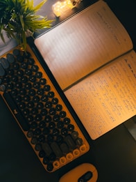 A dark workspace features a keyboard with round keycaps, an open notebook with handwritten notes, a small potted plant, and a lit decorative lamp. The overall setting suggests a cozy and focused writing or work environment.