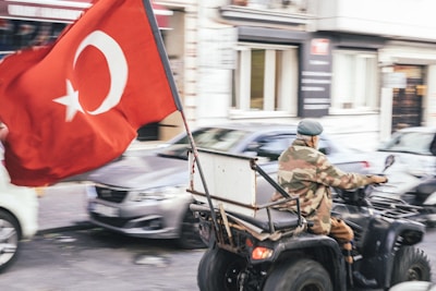 A person wearing camouflage attire rides a quad bike through a street, carrying a large Turkish flag. Several cars and buildings are visible in the background, giving the scene a sense of motion and urban environment.