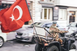 A person wearing camouflage attire rides a quad bike through a street, carrying a large Turkish flag. Several cars and buildings are visible in the background, giving the scene a sense of motion and urban environment.