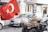 A person wearing camouflage attire rides a quad bike through a street, carrying a large Turkish flag. Several cars and buildings are visible in the background, giving the scene a sense of motion and urban environment.