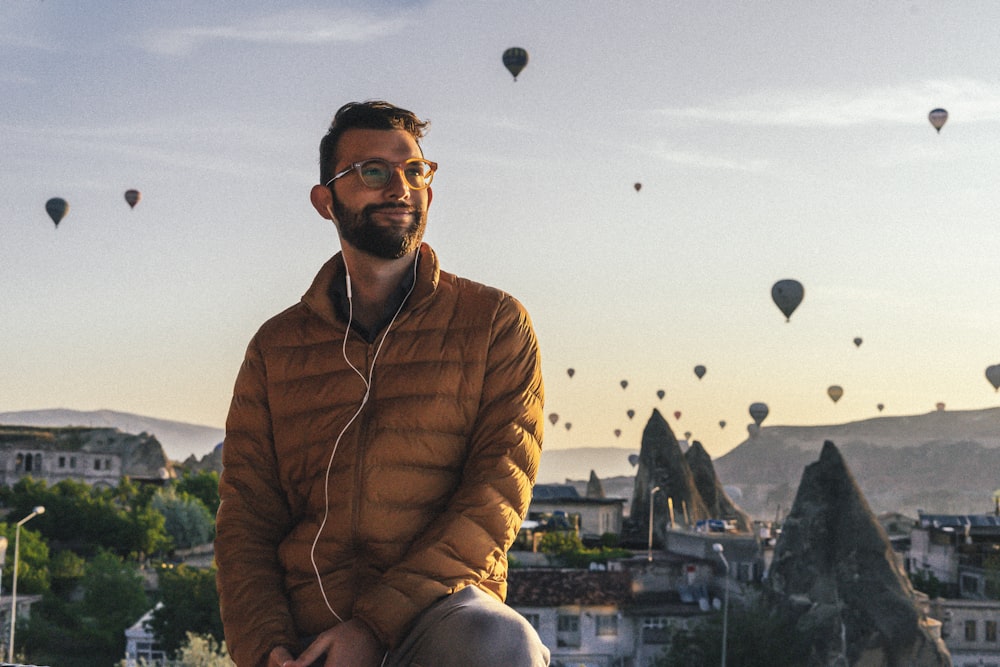 Un hombre sentado en una repisa con muchos globos aerostáticos en el cielo