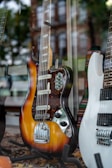 A vintage electric guitar with a sunburst finish and multiple control knobs is displayed prominently. Next to it is a modern white electric guitar. The setting is a shop window, as indicated by the reflections of a cityscape and trees.