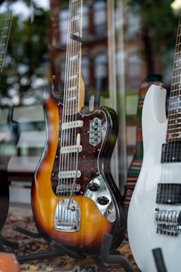 A vintage electric guitar with a sunburst finish and multiple control knobs is displayed prominently. Next to it is a modern white electric guitar. The setting is a shop window, as indicated by the reflections of a cityscape and trees.