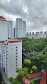 High-rise residential buildings with a backdrop of lush green trees. The buildings have a modern design, with white facades and red roofs. Overcast weather and cloudy skies create a serene atmosphere.