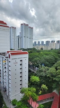High-rise residential buildings with a backdrop of lush green trees. The buildings have a modern design, with white facades and red roofs. Overcast weather and cloudy skies create a serene atmosphere.
