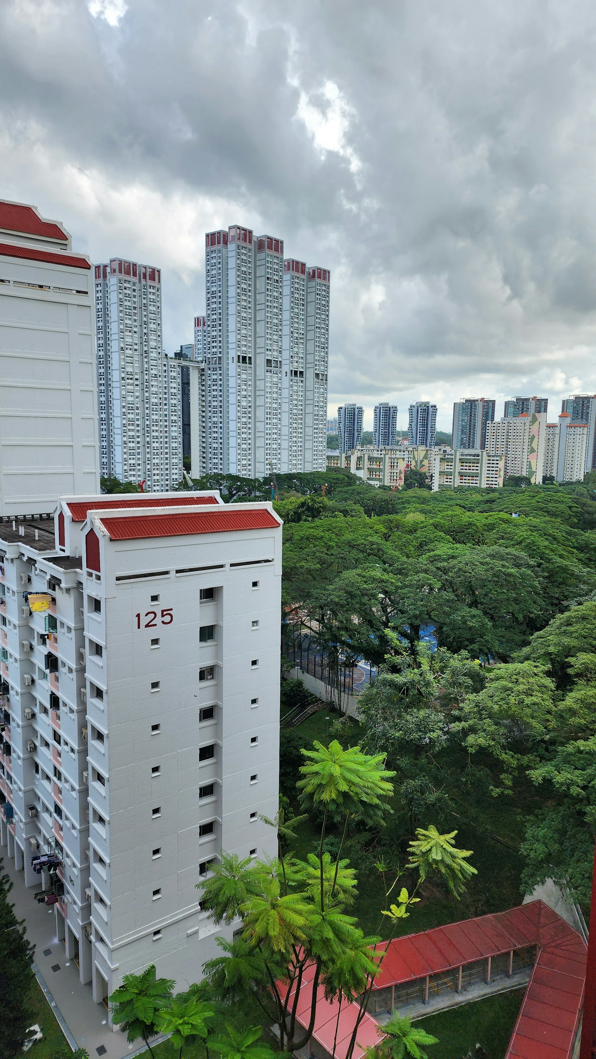 High-rise residential buildings with a backdrop of lush green trees. The buildings have a modern design, with white facades and red roofs. Overcast weather and cloudy skies create a serene atmosphere.