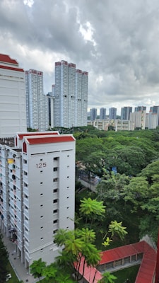 High-rise residential buildings with a backdrop of lush green trees. The buildings have a modern design, with white facades and red roofs. Overcast weather and cloudy skies create a serene atmosphere.