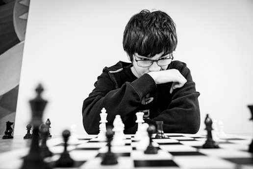 A person wearing glasses and a dark sweater is deeply focused on a chessboard. The black and white chess pieces are arranged in the middle of an intense game. The person appears to be contemplating their next move while resting their chin on their hand.