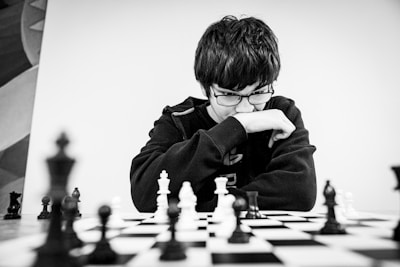 A person wearing glasses and a dark sweater is deeply focused on a chessboard. The black and white chess pieces are arranged in the middle of an intense game. The person appears to be contemplating their next move while resting their chin on their hand.