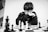 A person wearing glasses and a dark sweater is deeply focused on a chessboard. The black and white chess pieces are arranged in the middle of an intense game. The person appears to be contemplating their next move while resting their chin on their hand.