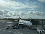 A passenger airplane is parked at an airport gate, with boarding equipment connected. Ground crew attend to luggage handling near the airplane, which features the branding 'Icelandair'. The sky is partly cloudy with rays of sunlight filtering through, creating a serene atmosphere. The runway and airport control tower are visible in the background.