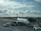 A passenger airplane is parked at an airport gate, with boarding equipment connected. Ground crew attend to luggage handling near the airplane, which features the branding 'Icelandair'. The sky is partly cloudy with rays of sunlight filtering through, creating a serene atmosphere. The runway and airport control tower are visible in the background.