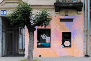 The storefront features a vibrant, colorful mural with bubble patterns painted in shades of pink, purple, and orange. Visible are a tree with lush green leaves, a sign with the number 55 on the wall, and a gate. The shop appears to be named 'Bubbles' with signage on a window and door.