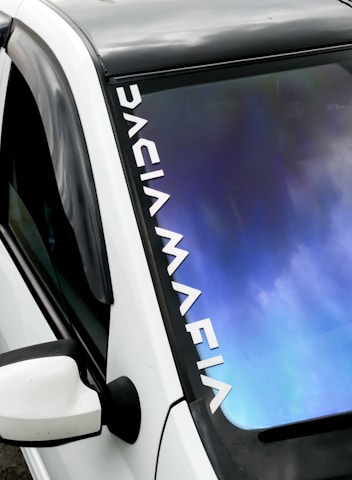 A close-up view of a car windshield featuring a reflective surface with a gradient of blue and purple tones. The car is white with a side mirror and a black roof. The words 'DACIA MAFIA' appear in bold white letters along the edge of the windshield.