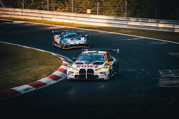 Two racing cars are taking a curved track on a race circuit. The leading car is branded with 'ROWE' and features a distinctive design, while another car follows closely behind. The track is lined with barriers and surrounded by greenery, indicating a professional racing environment.