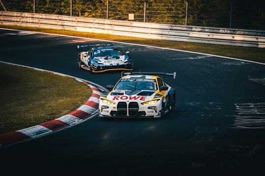 Two racing cars are taking a curved track on a race circuit. The leading car is branded with 'ROWE' and features a distinctive design, while another car follows closely behind. The track is lined with barriers and surrounded by greenery, indicating a professional racing environment.