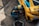 A mechanic in a blue and yellow racing suit is kneeling next to a yellow and blue race car, using a power tool to work on the wheel or tire. The car has various sponsor logos, including Michelin and Bilstein. The setting appears to be a pit stop area, with the ground displaying marks typical of race tracks.