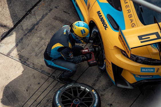 A mechanic in a blue and yellow racing suit is kneeling next to a yellow and blue race car, using a power tool to work on the wheel or tire. The car has various sponsor logos, including Michelin and Bilstein. The setting appears to be a pit stop area, with the ground displaying marks typical of race tracks.