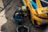 A mechanic in a blue and yellow racing suit is kneeling next to a yellow and blue race car, using a power tool to work on the wheel or tire. The car has various sponsor logos, including Michelin and Bilstein. The setting appears to be a pit stop area, with the ground displaying marks typical of race tracks.
