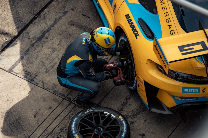 A mechanic in a blue and yellow racing suit is kneeling next to a yellow and blue race car, using a power tool to work on the wheel or tire. The car has various sponsor logos, including Michelin and Bilstein. The setting appears to be a pit stop area, with the ground displaying marks typical of race tracks.