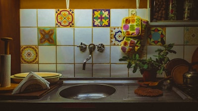 A kitchen sink area is adorned with colorful, patterned tiles. Hanging above the sink is a decorative oven mitt, and a small plant sits beside it on the countertop. Dishes and napkins are neatly arranged near a roll of paper towels. The ambient light creates a cozy and warm atmosphere.