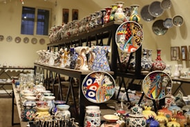 An array of intricately designed ceramic vases, plates, and pottery with vibrant patterns and colors is displayed on shelves in a room. The collection features various styles with detailed motifs and artistic designs, creating a visually rich scene.