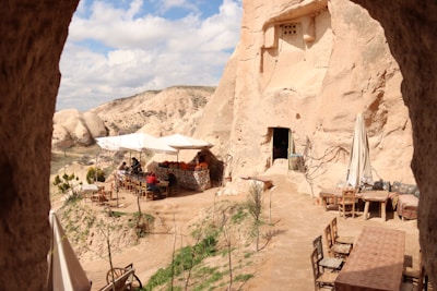 A rustic outdoor cafe set against a backdrop of rocky, sandy-colored cliffs. Several tables and wooden chairs are arranged on a dirt patio, with one table covered by a colorful tablecloth and a white umbrella. People are seated, enjoying the surroundings, with the entrance to a cave dwelling visible nearby.