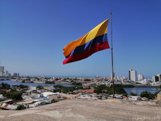 A large flag with yellow, blue, and red horizontal stripes is flying on a flagpole against a clear blue sky. Below, a cityscape with modern buildings, a body of water, and residential areas is visible, conveying a sense of openness and freedom.