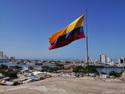 A large flag with yellow, blue, and red horizontal stripes is flying on a flagpole against a clear blue sky. Below, a cityscape with modern buildings, a body of water, and residential areas is visible, conveying a sense of openness and freedom.