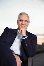 An individual with short gray hair and glasses is seated, wearing a formal dark suit and a white shirt. Their posture is relaxed with one hand resting on their chin, conveying a sense of confidence and ease. The background appears to be an outdoor setting with a blurred view of a cityscape at dusk, adding a serene atmosphere to the image.